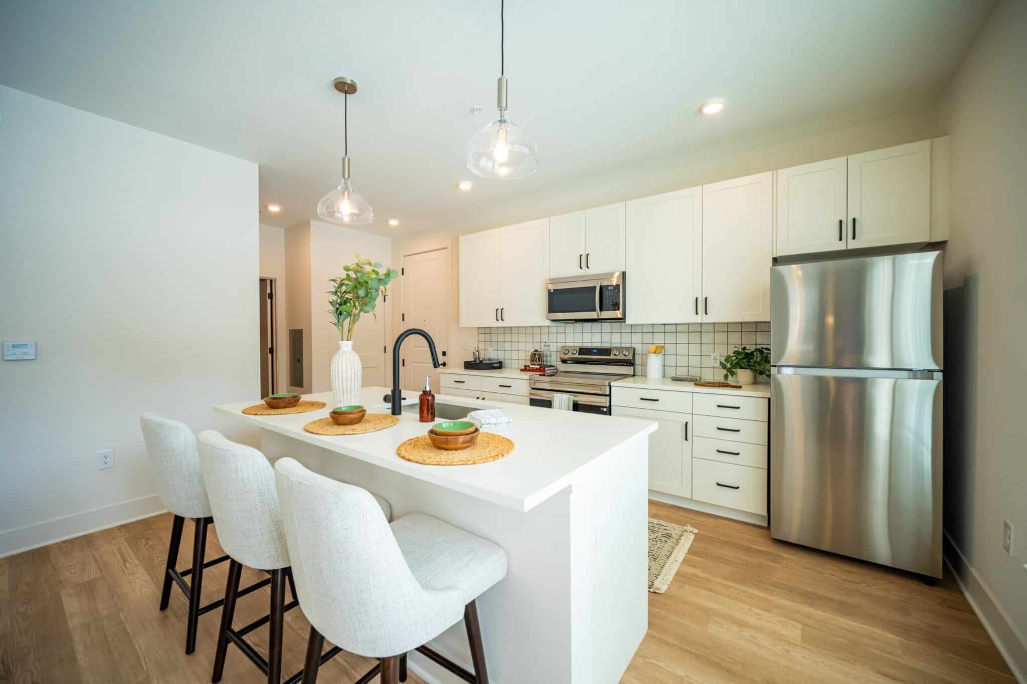 Modern kitchen with white cabinets, stainless steel appliances, an island with three upholstered stools, and pendant lights above. A vase with greenery sits on the island.
