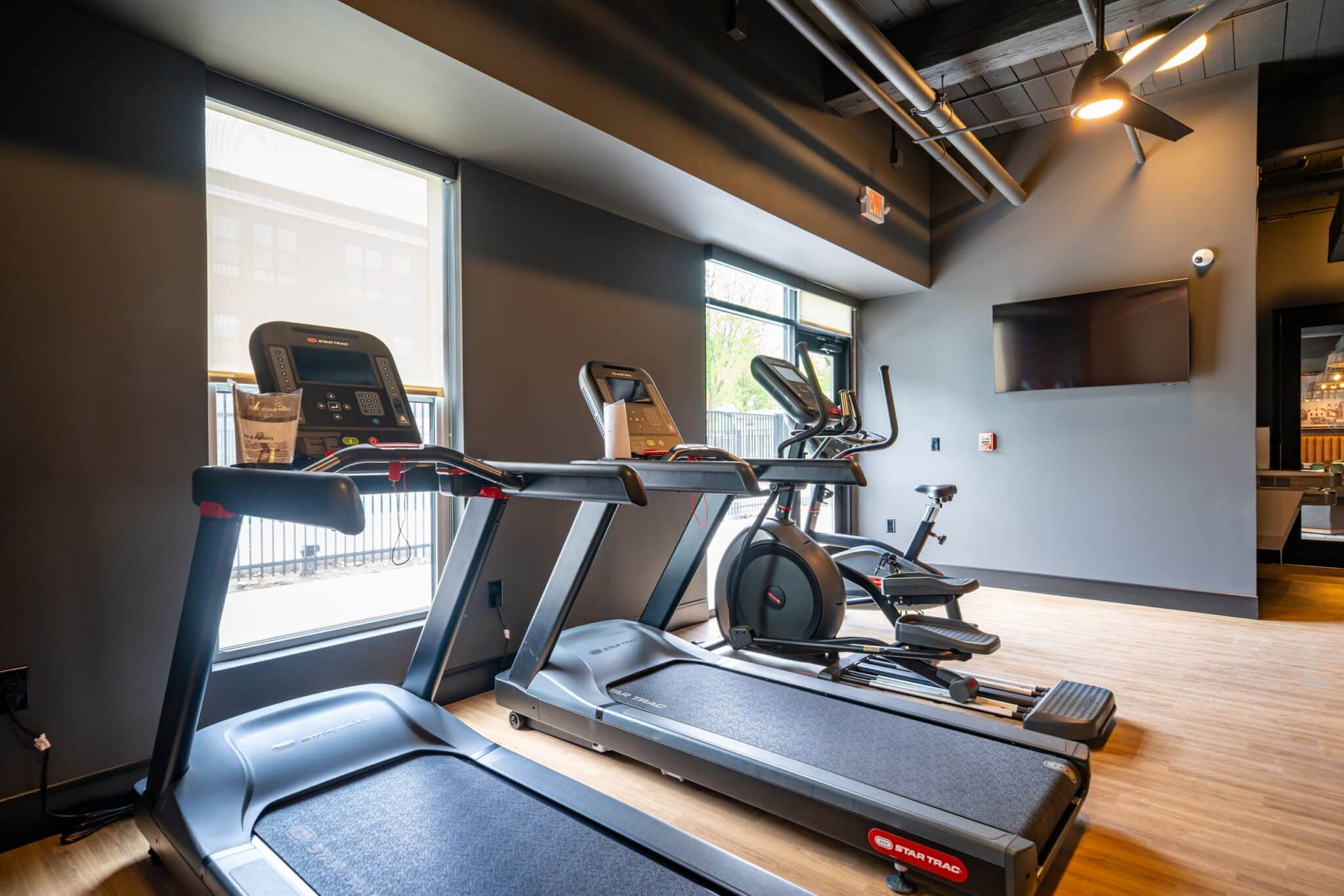 Modern gym room with two treadmills, an elliptical machine, and a stationary bike facing large windows and a wall-mounted TV.