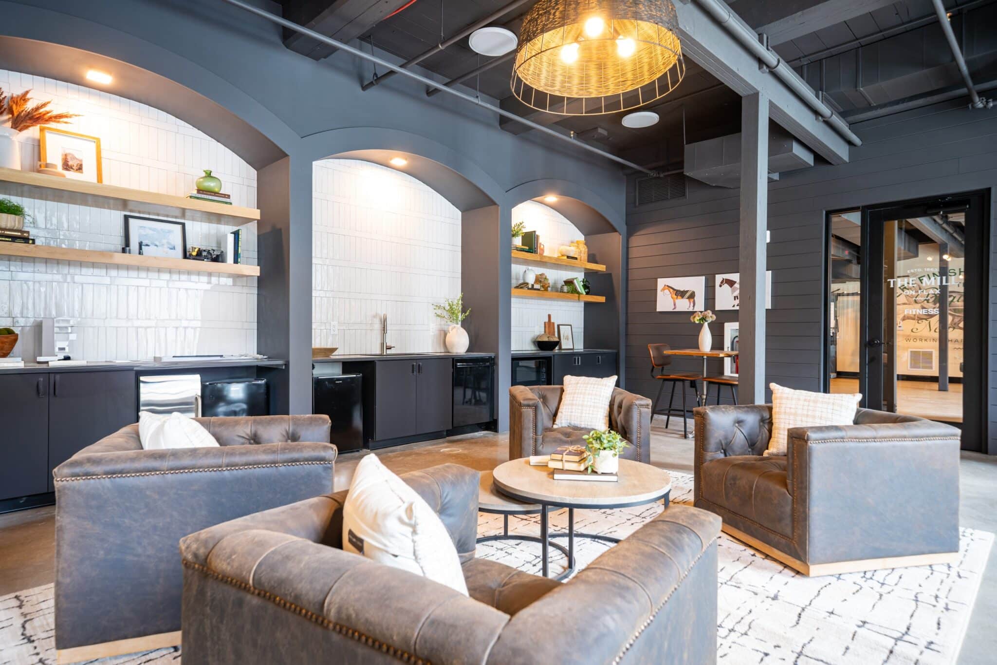 Modern lounge area with four leather armchairs, a round coffee table, built-in shelves, and decorative accents under archways; warm lighting and neutral tones throughout.