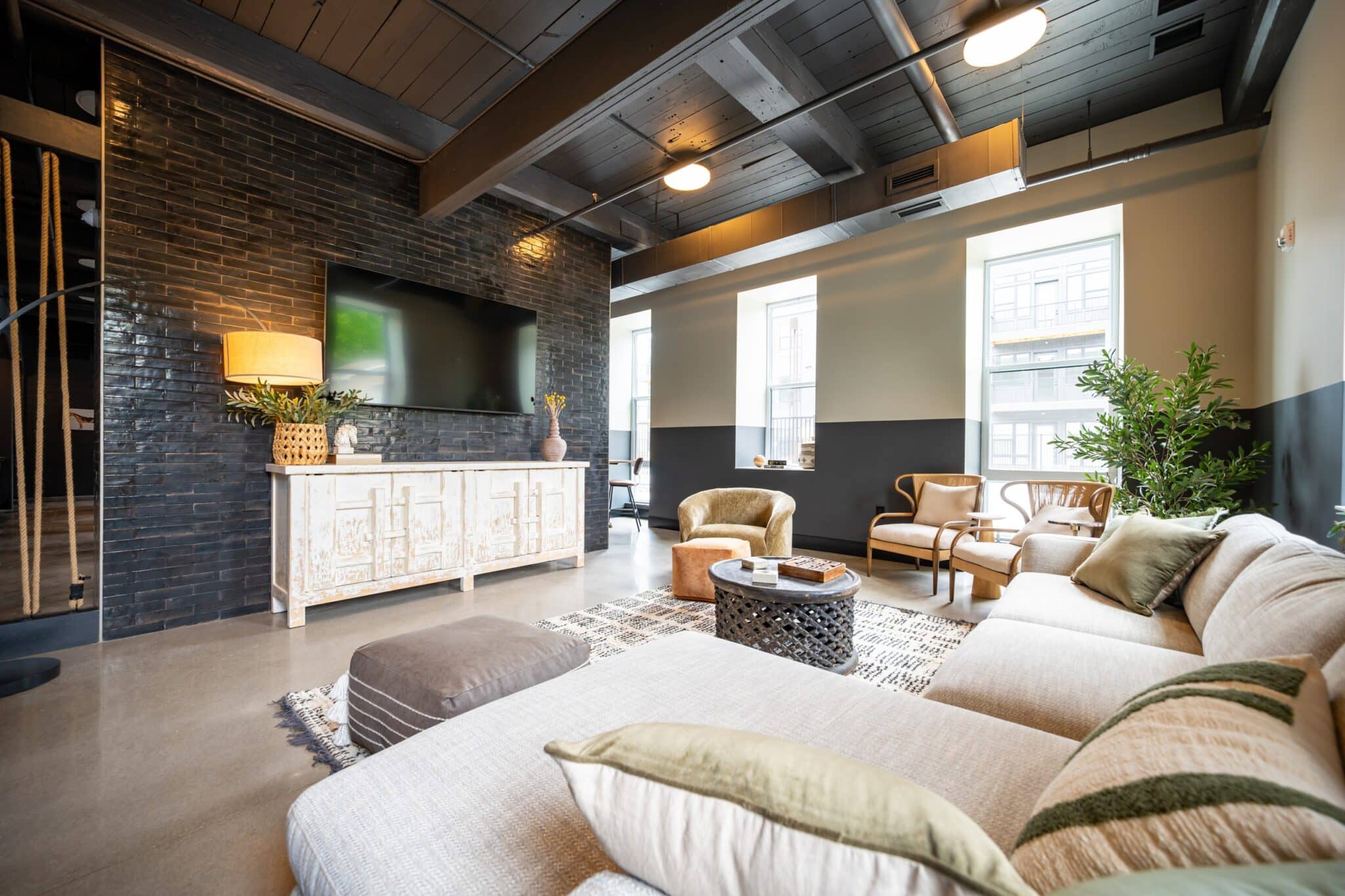 Modern living room with a sectional sofa, armchairs, a black brick accent wall with a mounted TV, a white console, large windows, and indoor plants.
