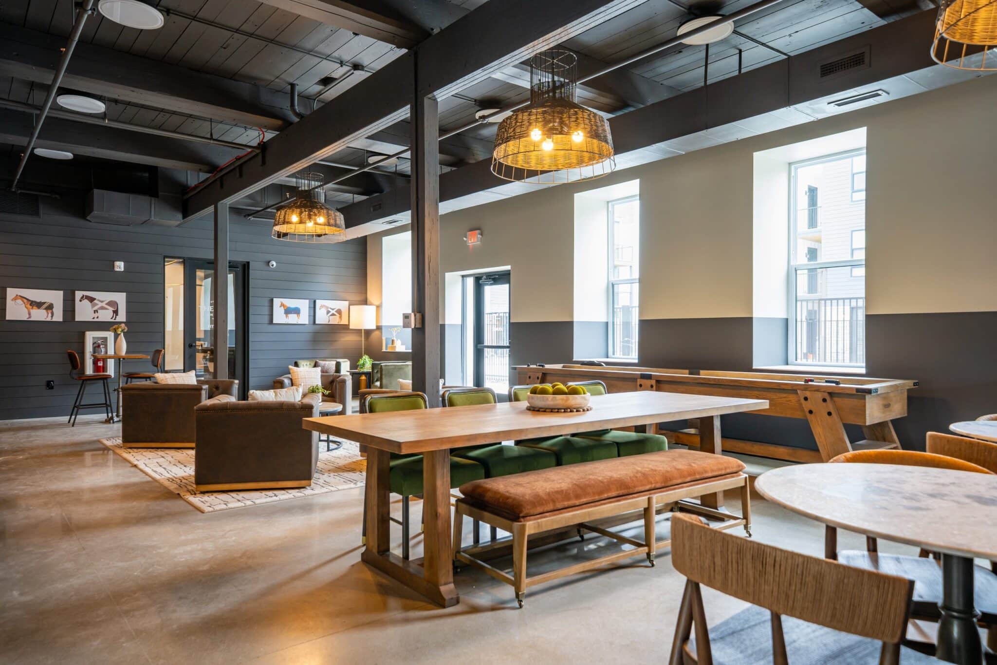 Modern communal lounge with wooden tables, bench seating, armchairs, wall art, foosball table, and large windows letting in natural light.