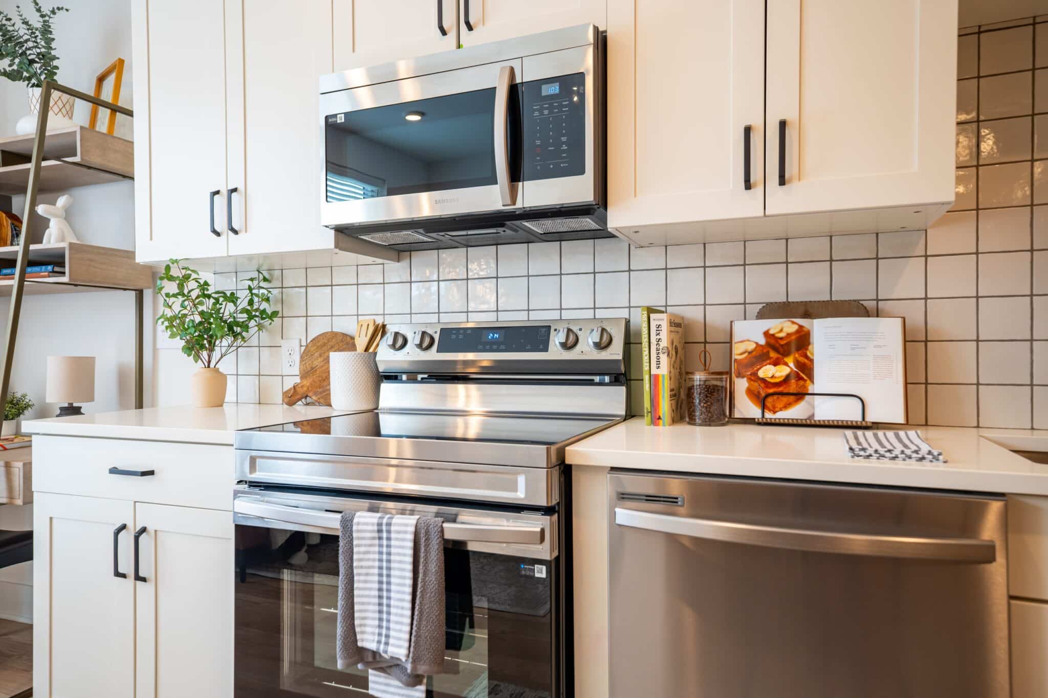 Modern kitchen with white cabinets, stainless steel appliances, a stove with a towel hanging, an open cookbook, and a potted plant on the counter.