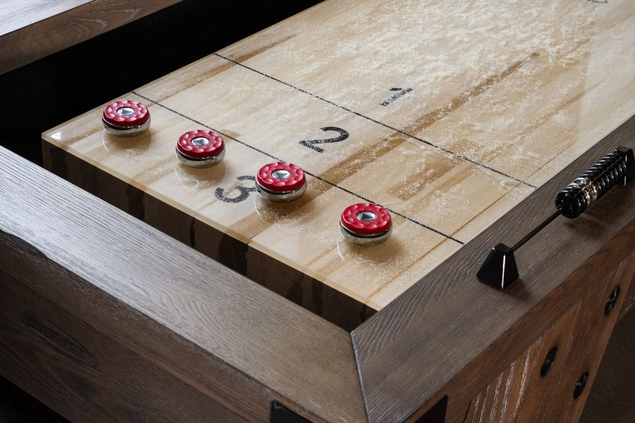 A close-up of a shuffleboard table by the old mill on flax, with four red pucks resting on numbered sections, covered in a light dusting of shuffleboard powder.