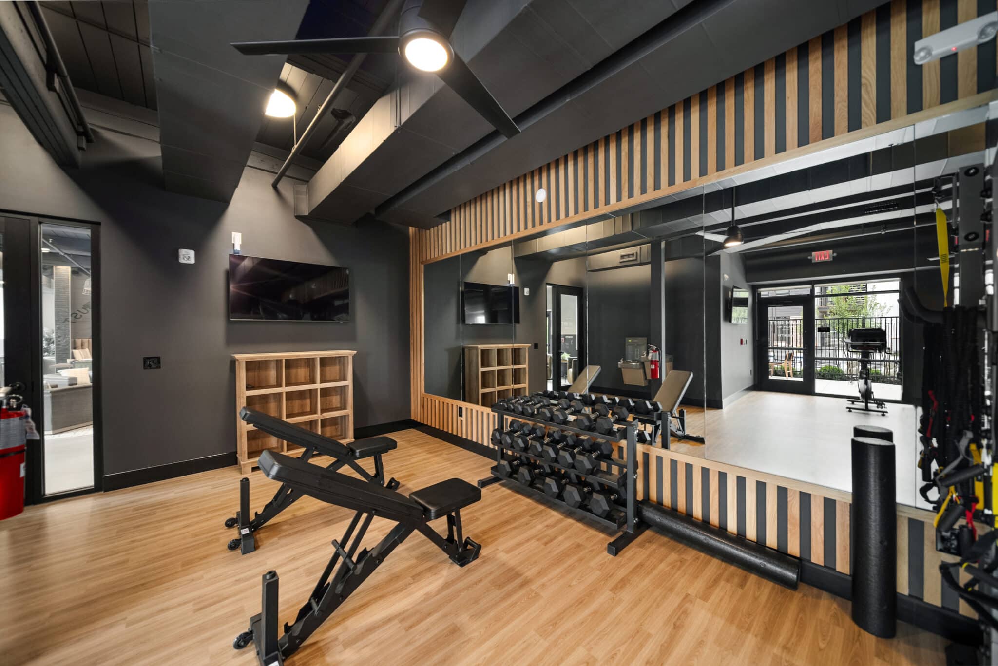A modern gym room with wooden floors, free weights on racks, adjustable benches, a large wall mirror, a TV, and floor-to-ceiling windows letting in natural light at Mill on Flax.