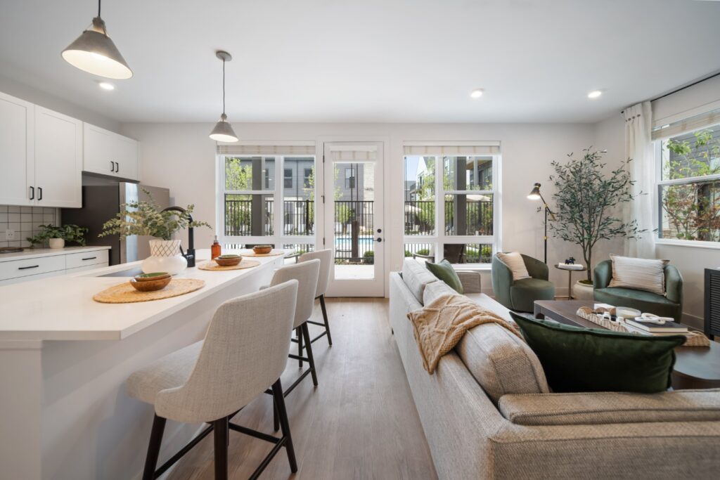 Modern open-plan kitchen and living room in the Mill on Flax style, featuring neutral decor, bar stools at the counter, a gray sofa with green cushions, and large windows that fill the space with natural light.
