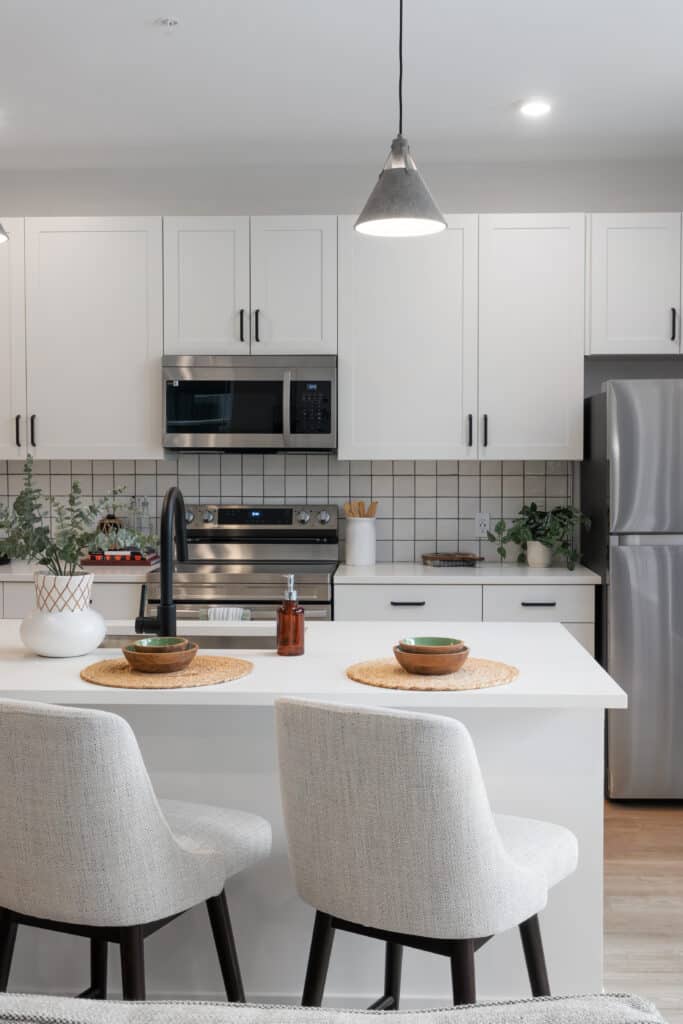 Experience urban convenience at The Mill on Flax in this modern kitchen featuring white cabinets, stainless steel appliances, and an island with two upholstered chairs. Lush plants and stylish kitchen items decorate the counters.