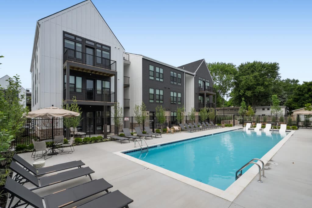 Outdoor swimming pool with lounge chairs and umbrellas beside the modern three-story Mill on Flax apartment building, surrounded by trees and landscaping.