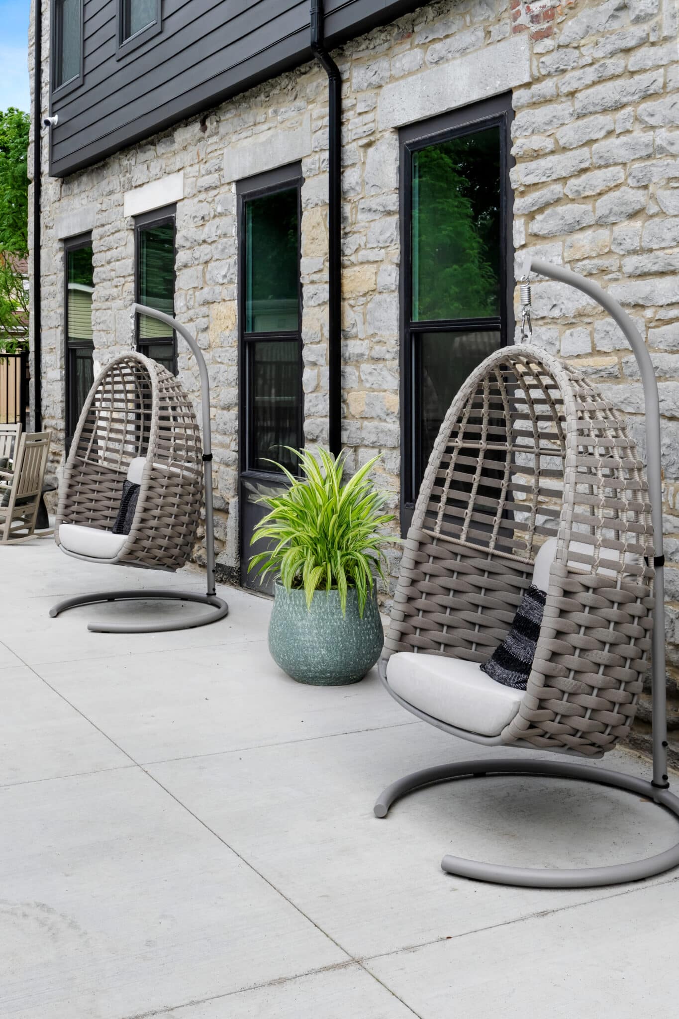 Two hanging wicker chairs with cushions sit on a concrete patio beside a stone building, just steps from the historic mill on Flax, with a green potted plant placed between them.