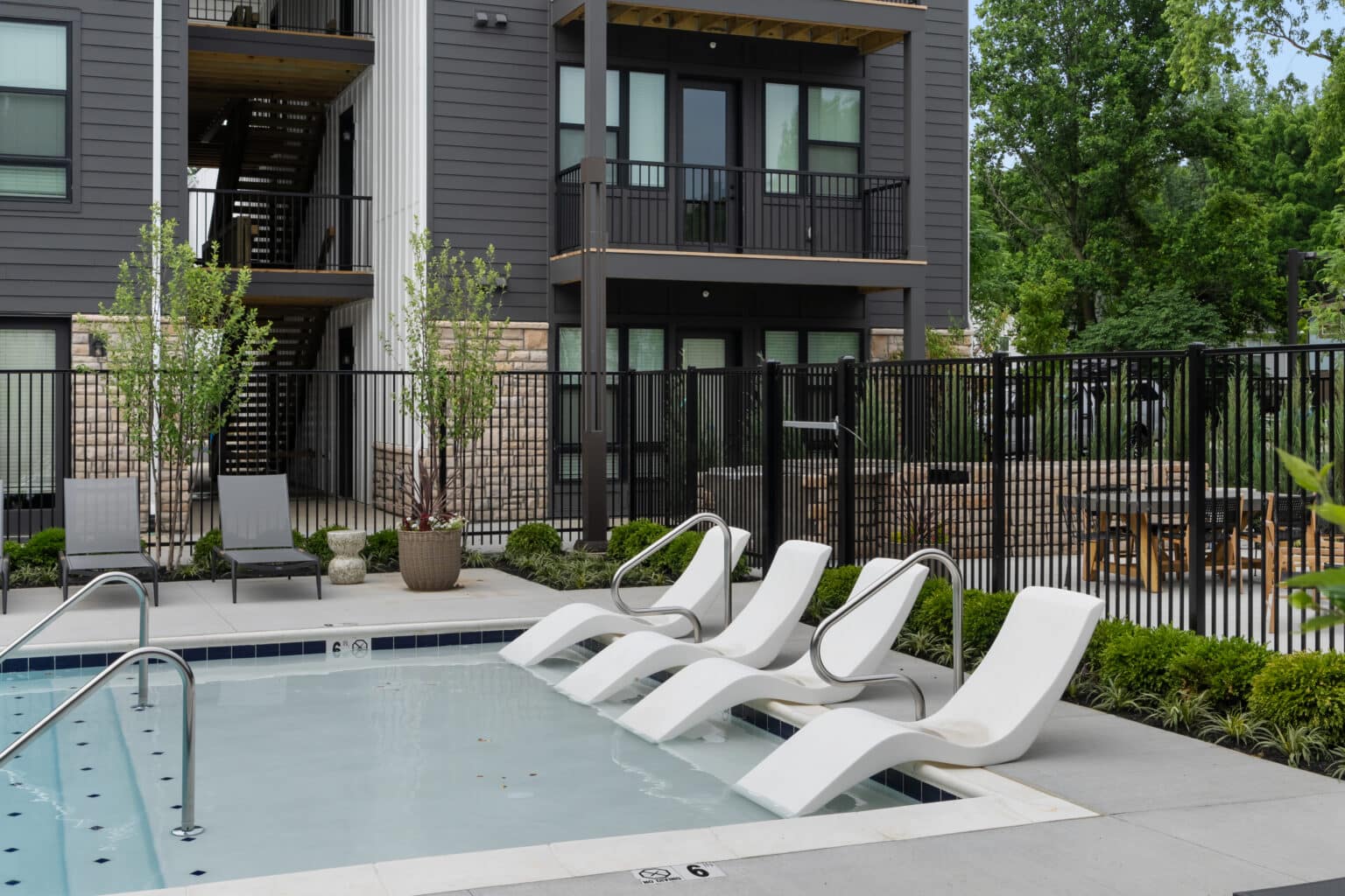 Three white lounge chairs rest in the shallow end of an outdoor pool near a modern apartment building with balconies—Experience Urban Convenience at The Mill on Flax.
