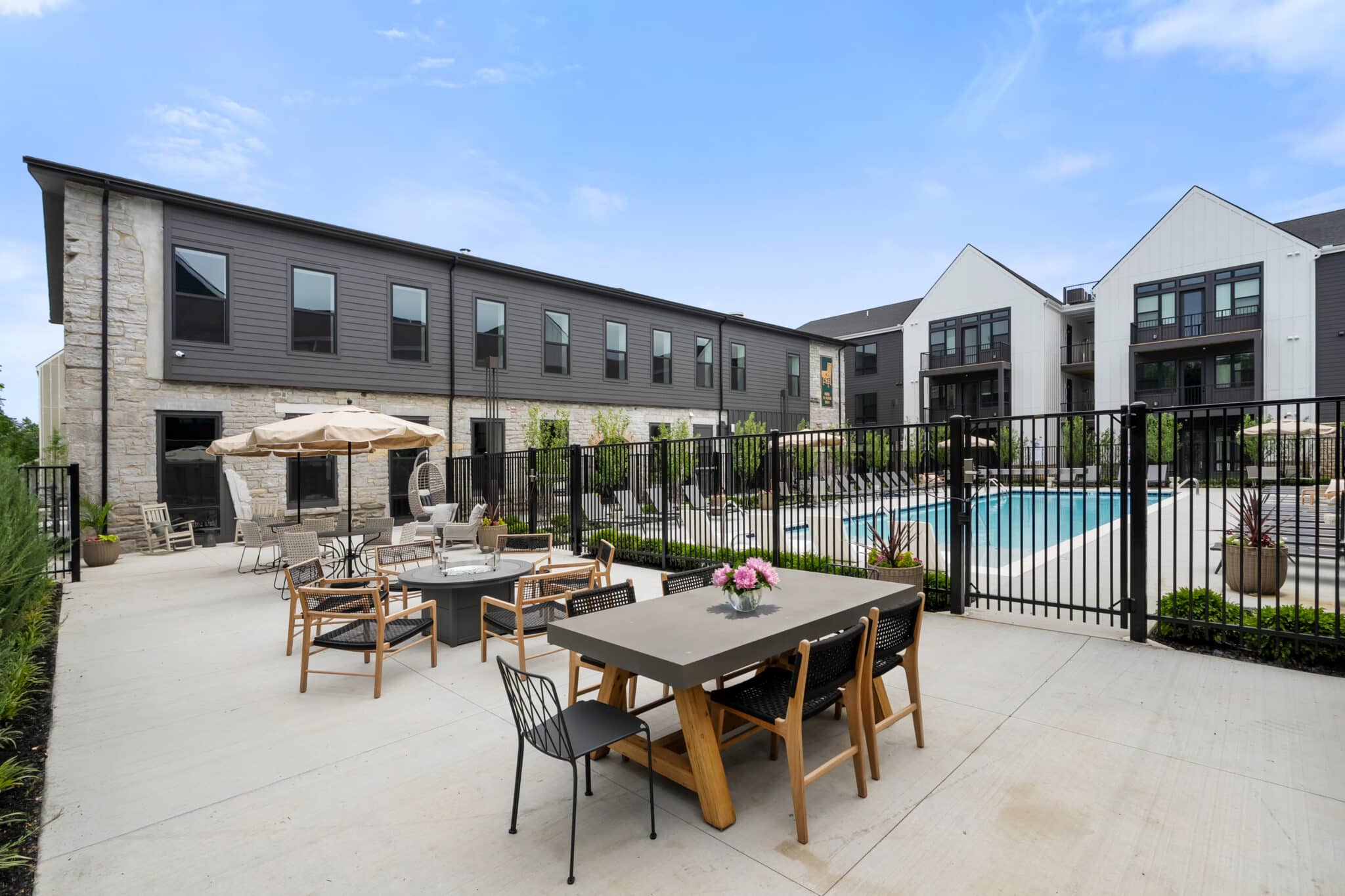 Modern apartment complex courtyard at Mill on Flax, featuring an outdoor dining table, fire pit seating area, and a fenced-in swimming pool under a clear blue sky.