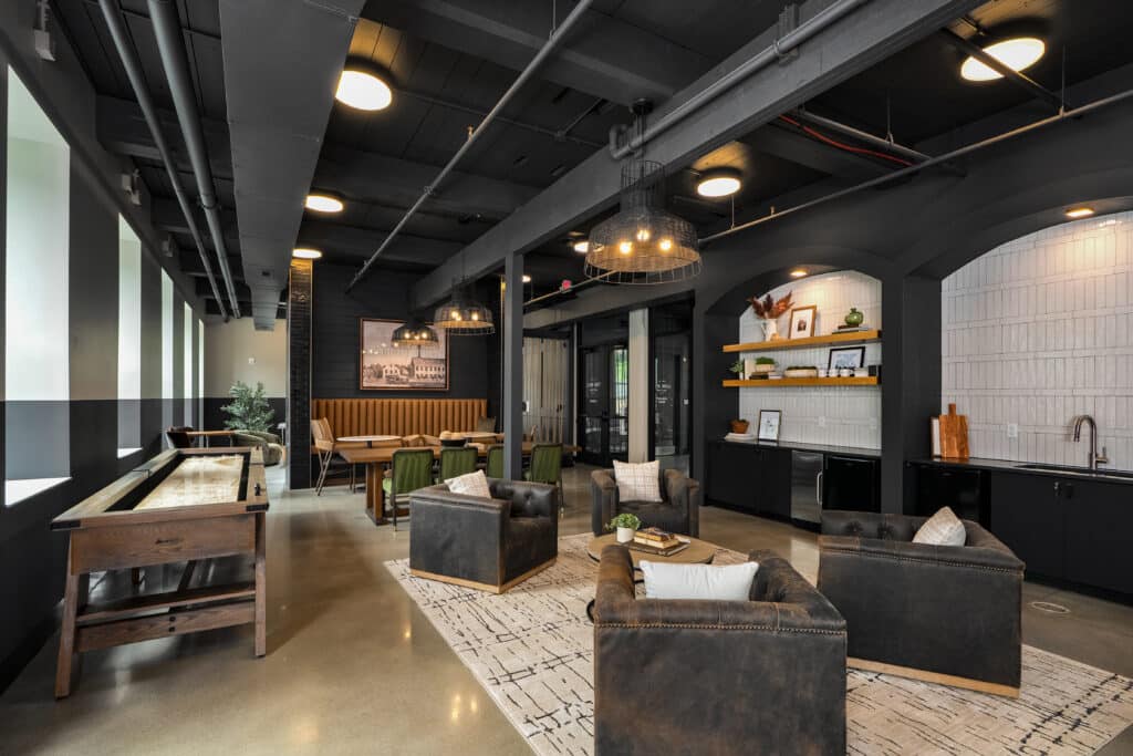 Modern lounge area with leather armchairs, a wooden shuffleboard table, dining nook, and open shelving evokes the character of a mill on flax, featuring an industrial-style ceiling with exposed pipes and lighting.