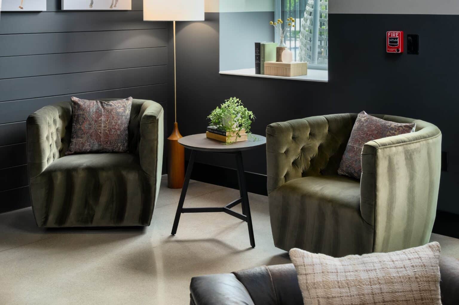 Two green velvet armchairs with patterned cushions sit around a small round black table with a potted plant, creating a stylish corner inspired by Mill on Flax, in a modern room with grey walls and a floor lamp.