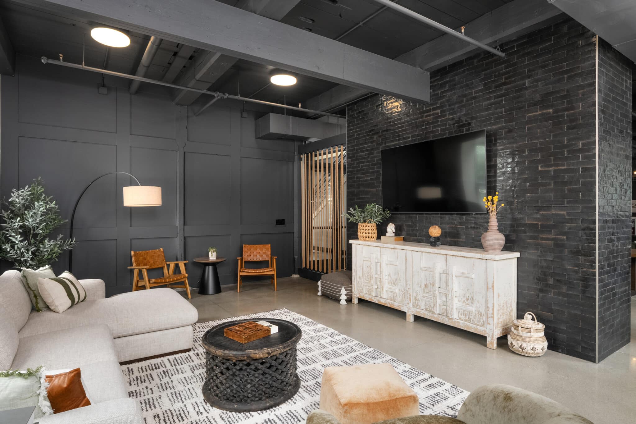 Modern living room with dark walls, mounted TV, white sideboard, beige sofa, woven coffee table, and leather chairs. Decorative plants add freshness. Exposed ceiling beams and a concrete floor recall the charm of a converted mill on flax.