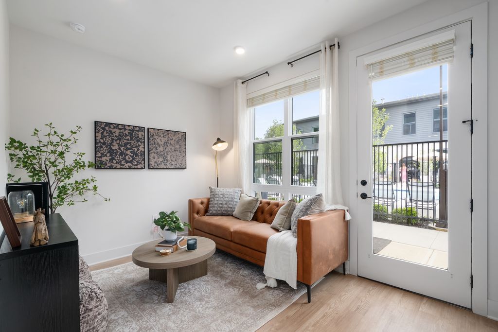 A modern studio apartment living room with a brown leather sofa, round wooden coffee table, rug, black cabinet, potted plants, and large windows letting in natural light.