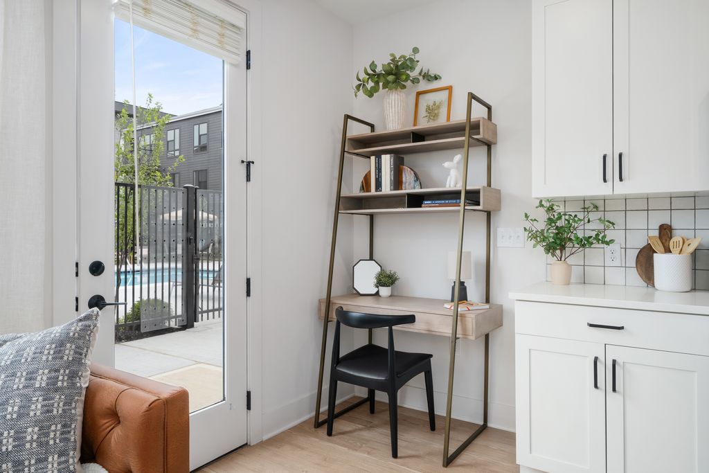 A cozy studio apartment features a small home office nook with a wooden desk, black chair, and shelves, tucked beside a white kitchen cabinet and a glass door that opens to a pool area outside.