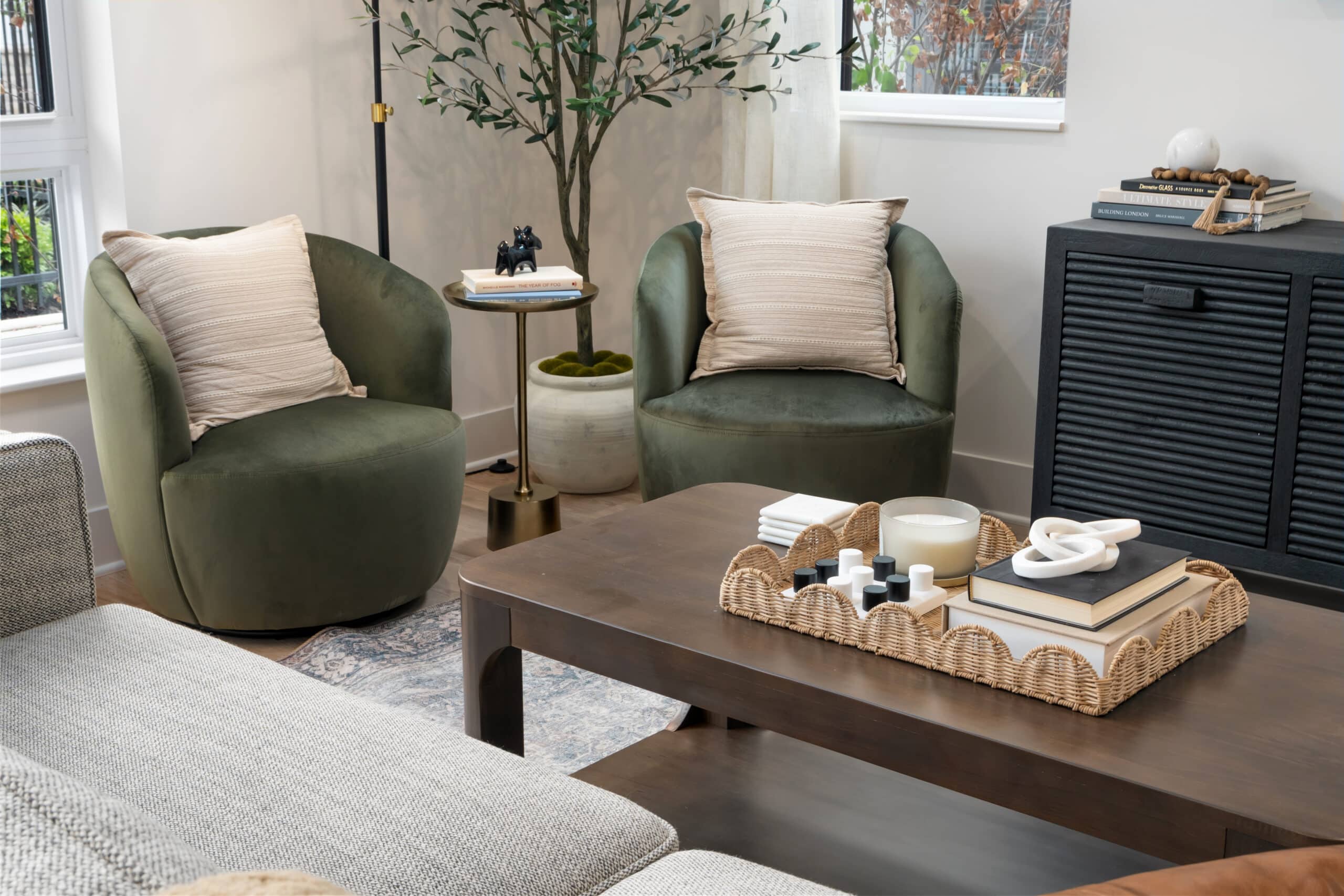 A modern studio apartment living room with two green armchairs, a wooden coffee table topped with a wicker tray, and decorative items arranged near a potted plant and cabinet.