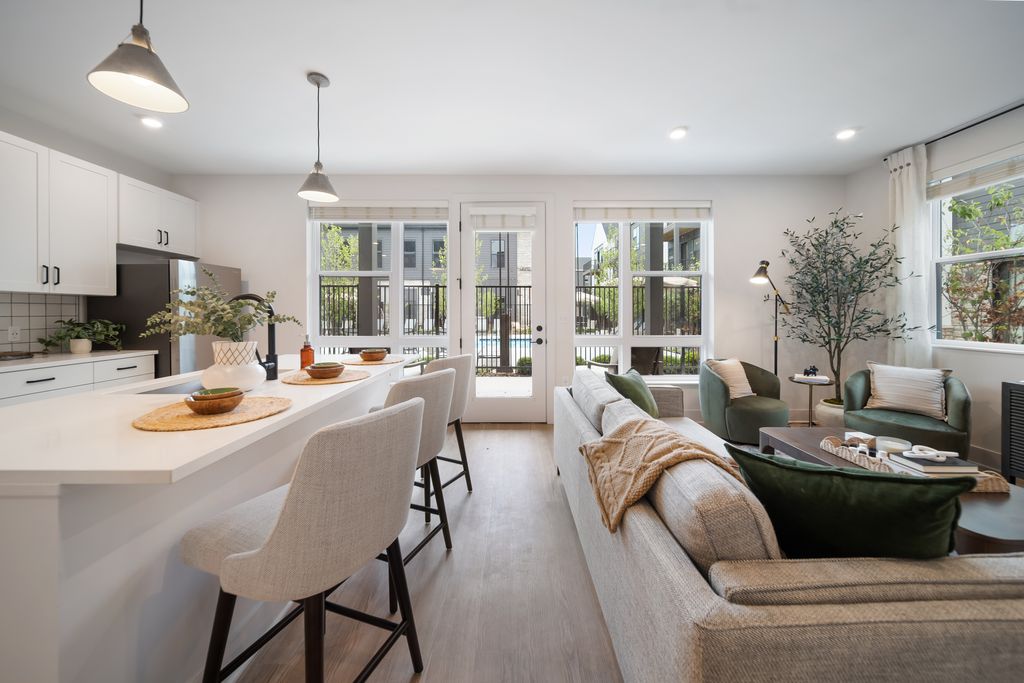Modern open-concept studio apartment with neutral decor, bar stools at a white island, a gray sofa, green chairs, large windows, and ample natural light.