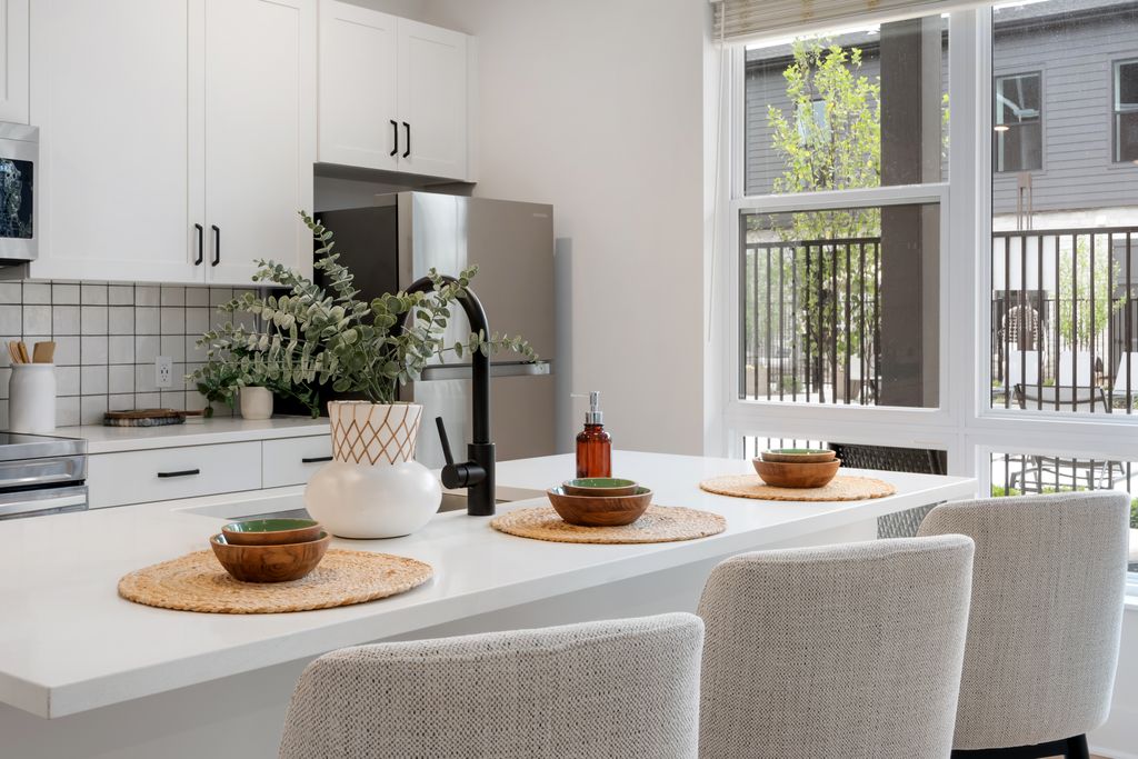 Modern kitchen in a studio apartment with white cabinets, a large island with three place settings, a vase with greenery, and a window letting in natural light.