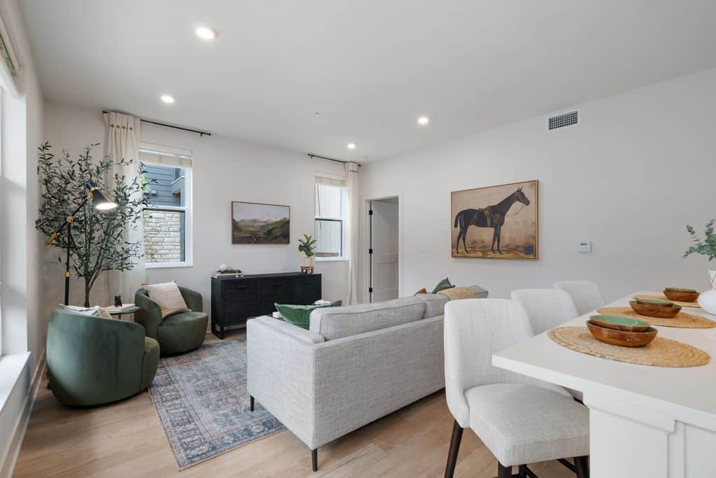 Modern studio apartment featuring a light gray sofa, green armchairs, wall art, and a dining counter with place settings. The space boasts wood flooring, indoor plants, and neutral decor for a stylish yet cozy ambiance.
