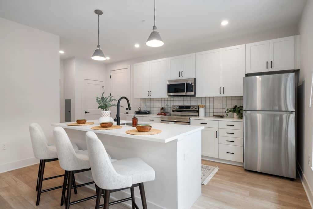 Modern studio apartment kitchen with white cabinets, stainless steel appliances, a large island with three upholstered bar stools, pendant lighting, and decorative place settings.