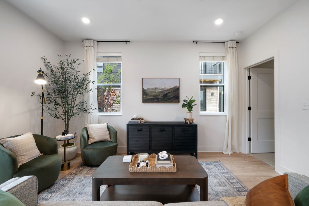 Modern studio apartment living room with neutral walls, two green chairs, a gray sofa, a wooden coffee table, black sideboard, plants, and a landscape painting between two windows with white curtains.