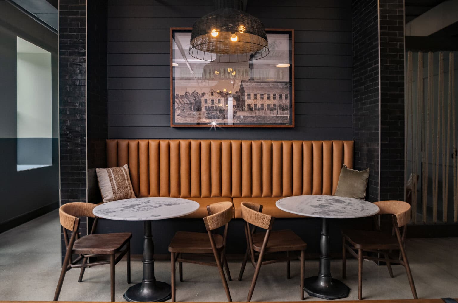A modern cafe seating area featuring a brown leather bench, two round marble tables, wooden chairs, and a framed cityscape photo on a dark paneled wall offers stylish design and comfortable amenities.