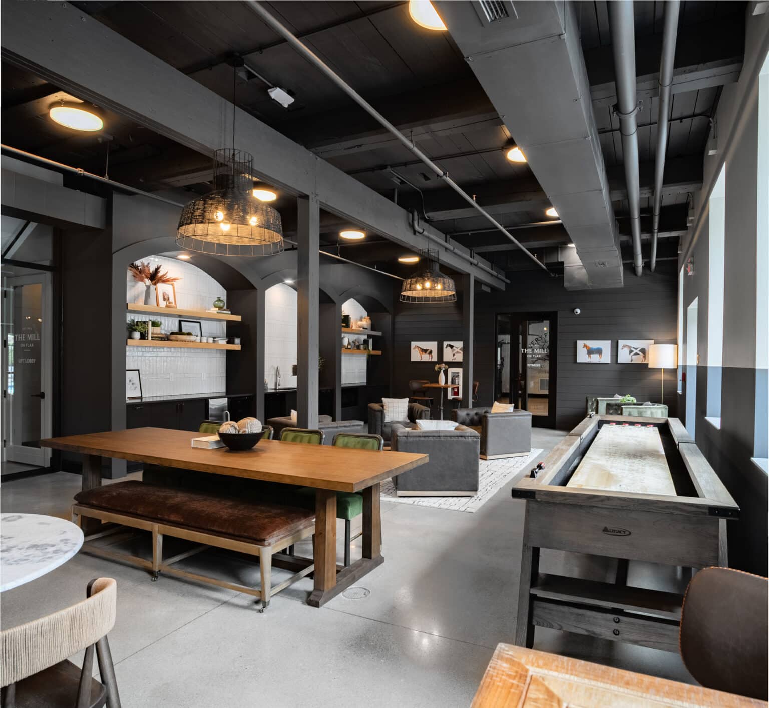 Modern lounge area with wooden tables, gray sofas, a shuffleboard table, open shelving, and framed art on dark walls under an exposed industrial ceiling—offering stylish amenities for relaxation and socializing.