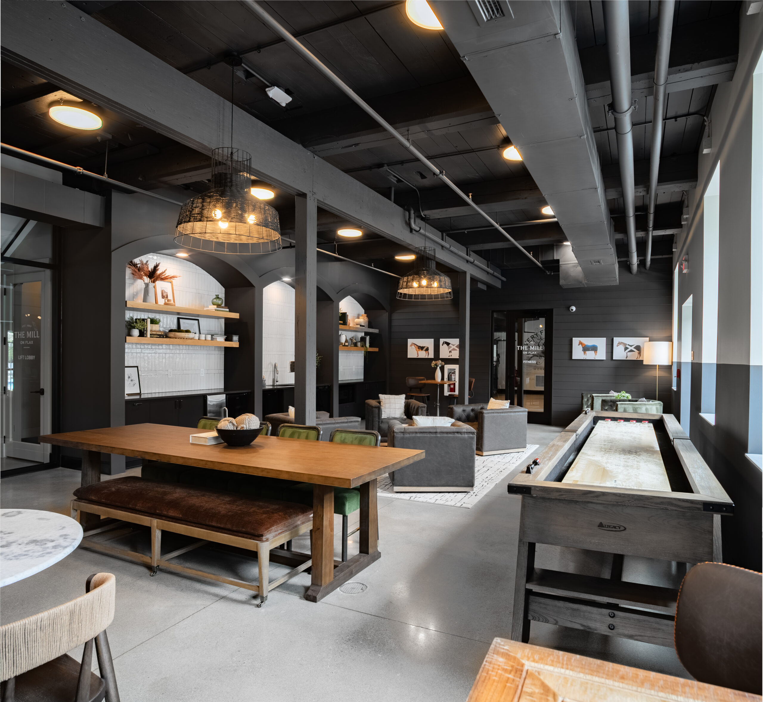 A modern lounge featuring dark walls, a wood dining table, gray seating area, shelves with decor, framed art, and excellent amenities like a shuffleboard table along the right side.