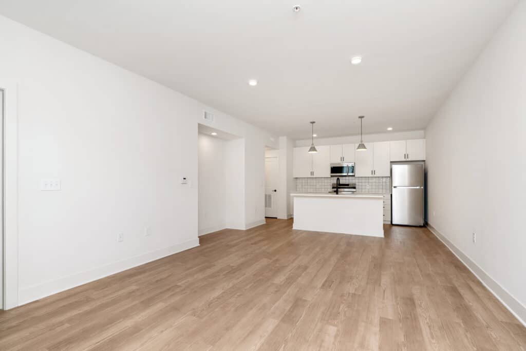 Modern empty one bedroom apartment with light wood flooring, white walls, recessed lighting, and an open kitchen featuring a center island, stainless steel appliances, and pendant lights.