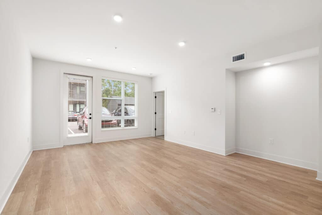 Unfurnished one bedroom apartment features light wood flooring, white walls, recessed lighting, a large window, and a glass door leading outside.