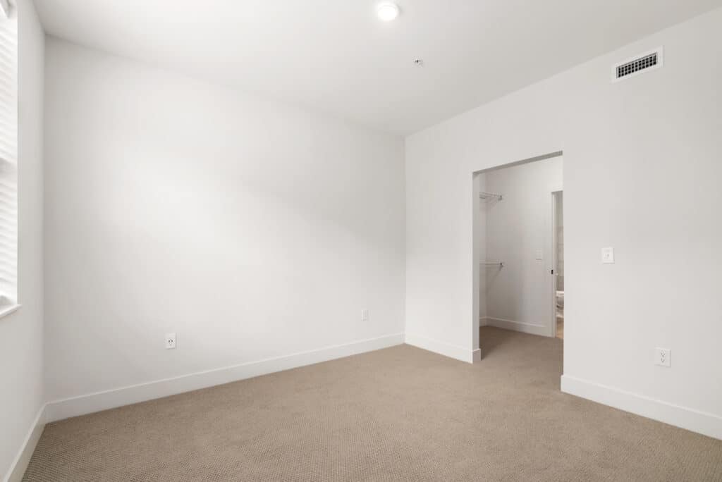 Empty room in a one bedroom apartment with beige carpet, white walls, a window on the left, and an open doorway leading to a closet and bathroom area. Ceiling light and air vent are visible.