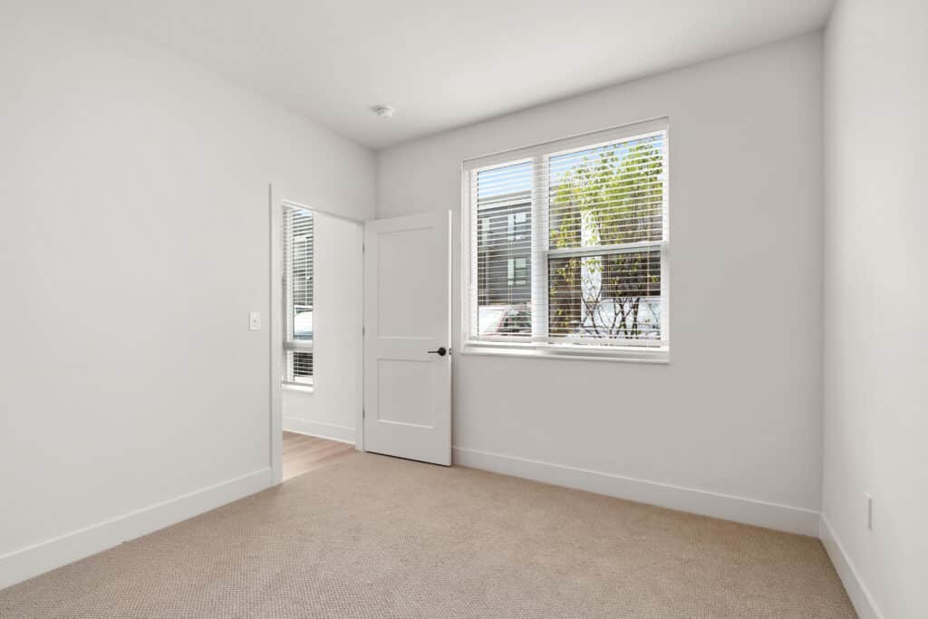 Empty room in a one bedroom apartment features beige carpet, white walls, a window with blinds, and an open door leading to another room with wooden flooring.
