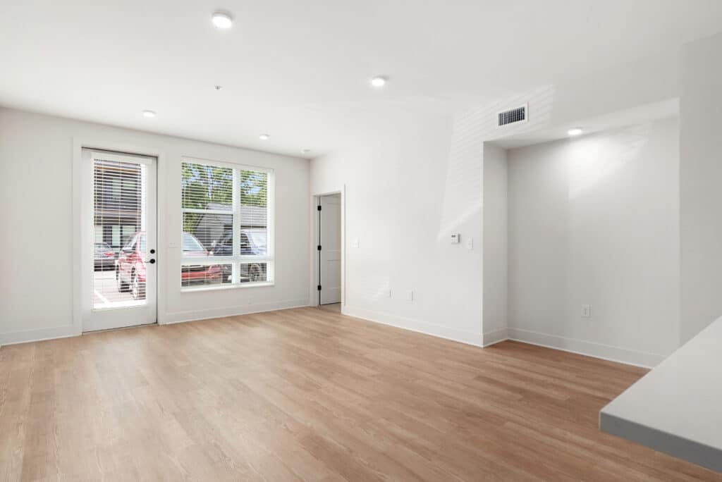 Empty, modern living room in a one bedroom apartment with light wood floors, white walls, recessed lighting, large windows, a door to the outside, and two interior doors.