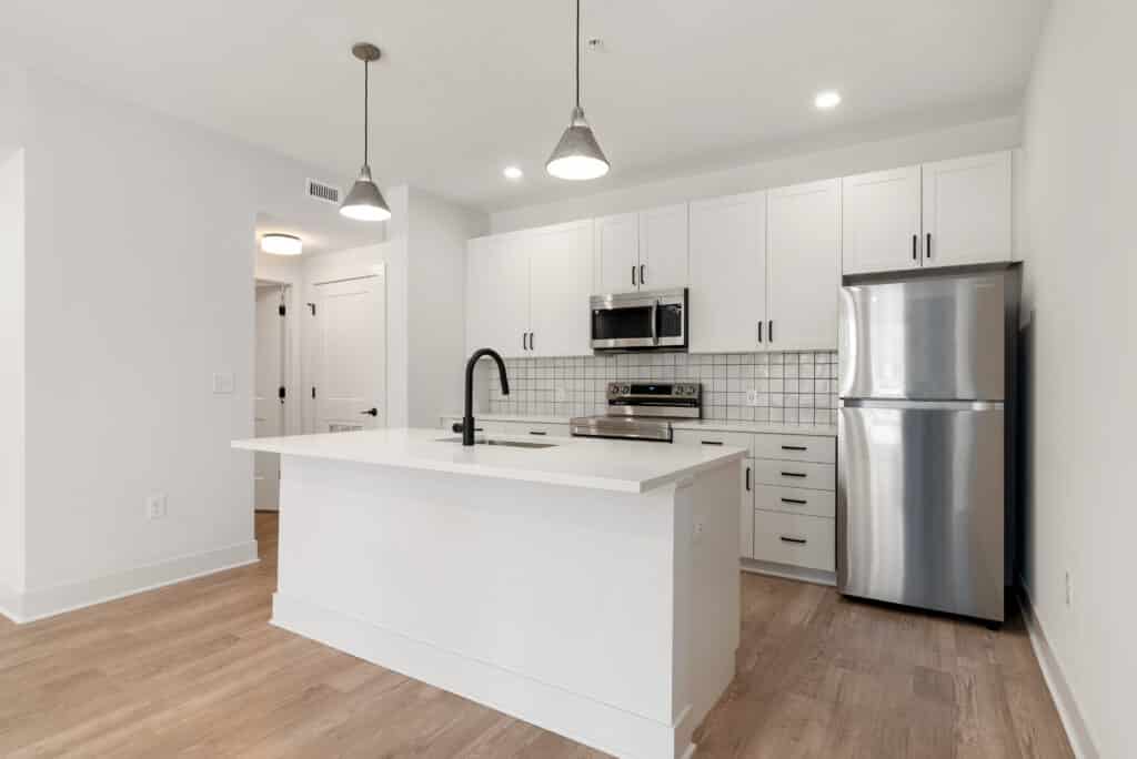 Modern kitchen in a One Bedroom Apartment featuring white cabinets, a stainless steel refrigerator and stove, a kitchen island with a sink, pendant lights, and light wood flooring.