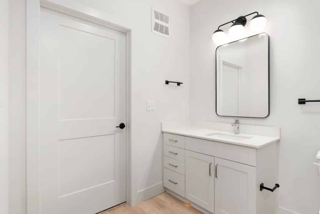 Modern bathroom in a One Bedroom Apartment with white walls, a rectangular mirror above a sleek white sink, vanity with drawers, black fixtures, and a crisp white door.