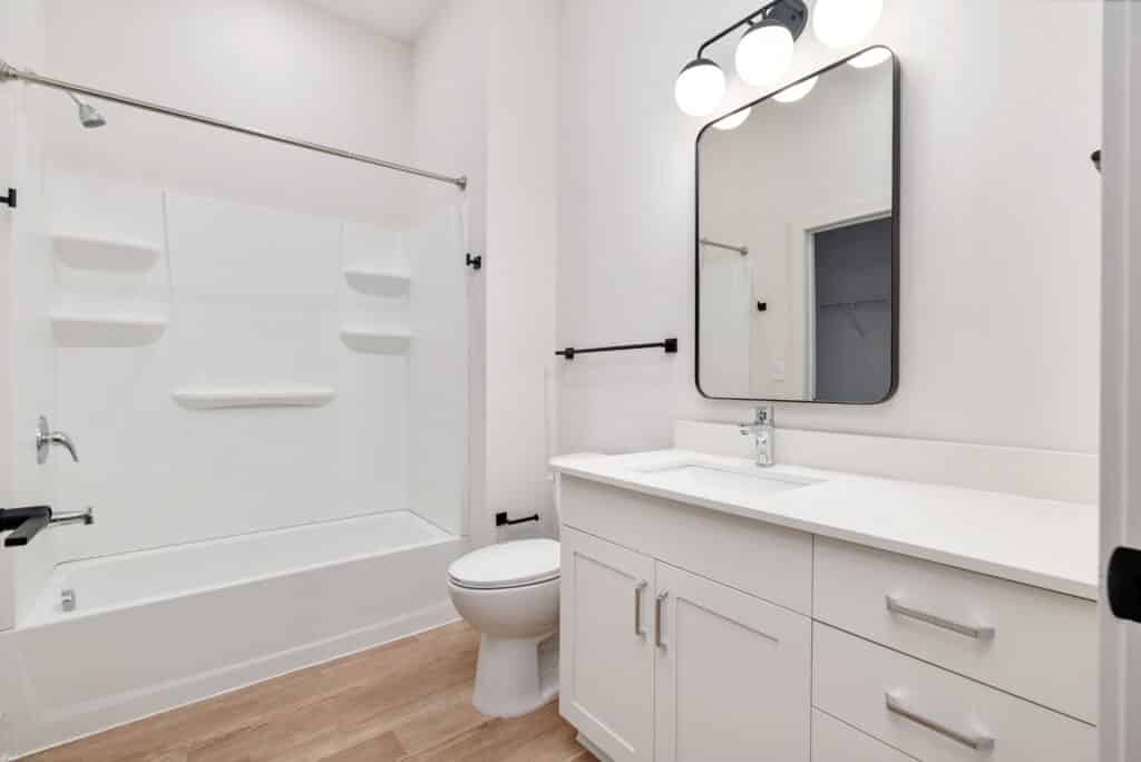Modern bathroom in a Two Bedroom Apartment with a white bathtub-shower combo, toilet, white vanity with sink, wall mirror, and overhead lights, featuring wood-style flooring and minimal decor.