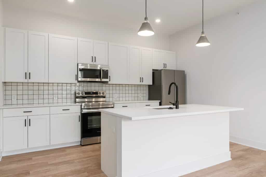 Modern kitchen in a two bedroom apartment with white cabinets, stainless steel appliances, a center island with sink, light wood flooring, and pendant lights.