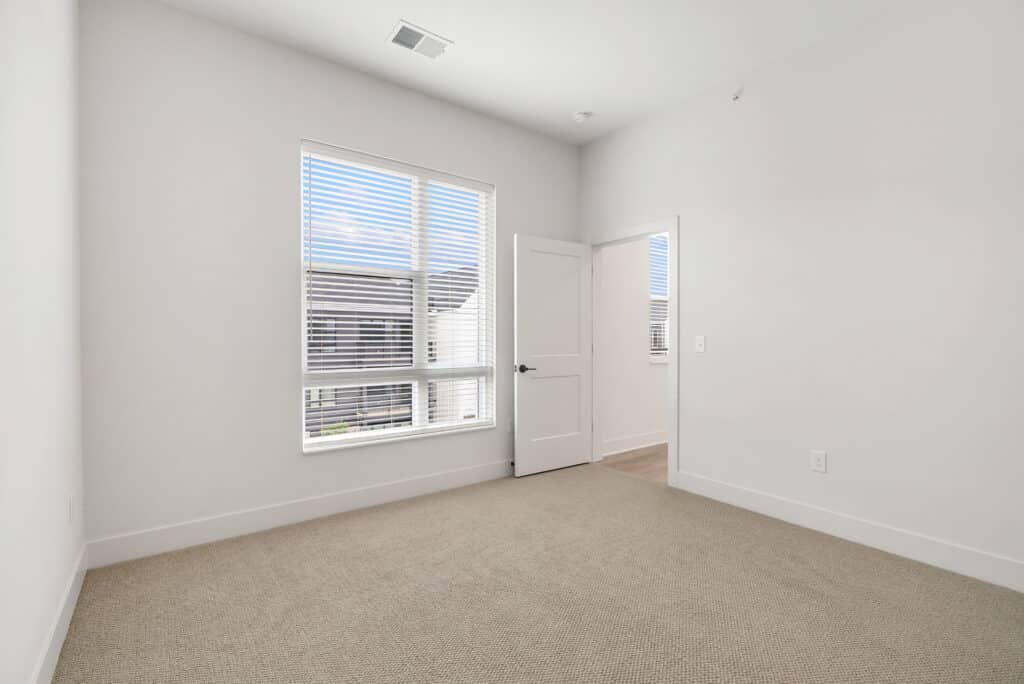 Empty room with beige carpet, white walls, a large window with blinds, and an open door leading to a hallway in this spacious two bedroom apartment.