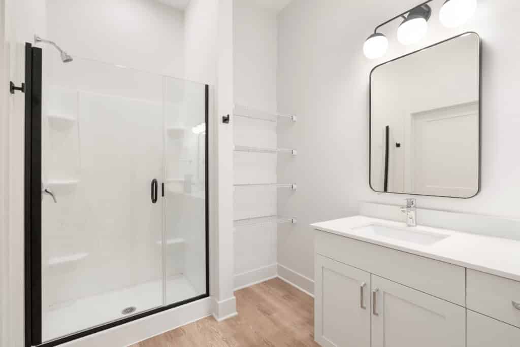Modern bathroom in a Two Bedroom Apartment featuring a glass shower, white vanity with mirror, wall-mounted light fixture, and wire shelving set against white walls and a light wood floor.