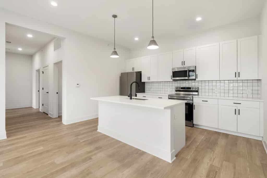 Modern kitchen in a two bedroom apartment featuring white cabinets, stainless steel appliances, tile backsplash, central island with black faucet, pendant lighting, and light wood flooring.