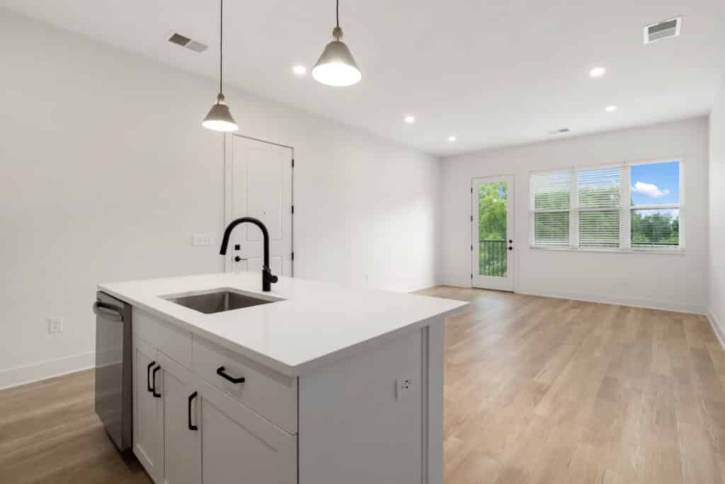 Modern, unfurnished two bedroom apartment featuring an open-plan kitchen and living area with white walls, wooden flooring, island sink, pendant lights, and large windows overlooking greenery.