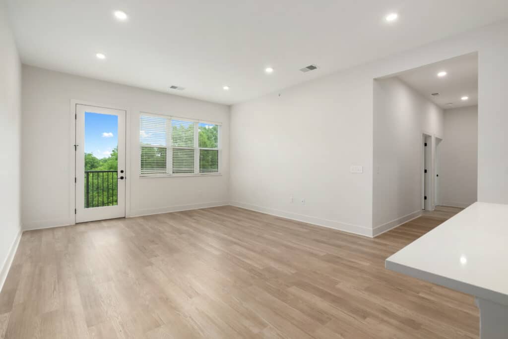 Empty, unfurnished living room in a two bedroom apartment with light wood floors, white walls, large windows, a glass door to a balcony, and recessed ceiling lights.