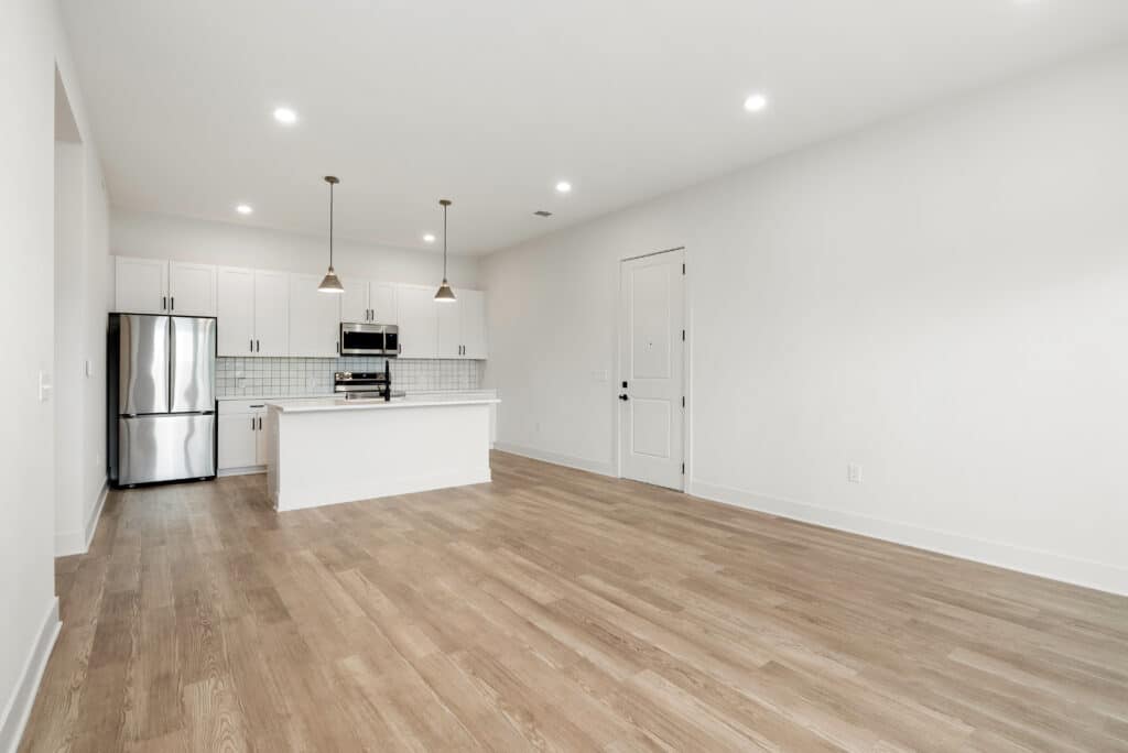 Empty modern two bedroom apartment interior with an open kitchen, stainless steel appliances, white cabinets, pendant lights, and light wood flooring.