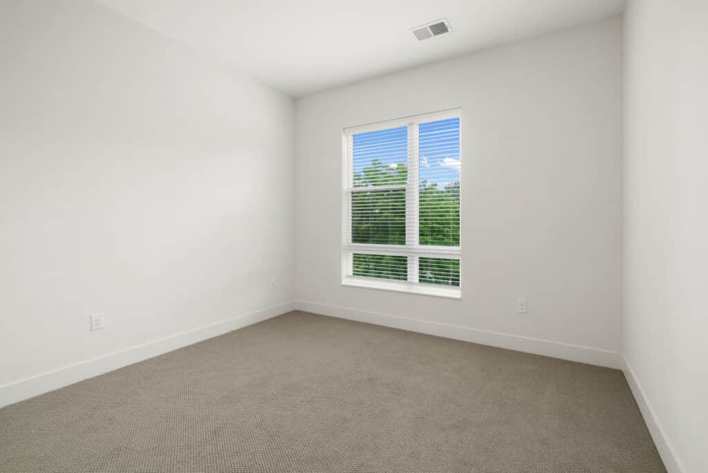 Empty room with white walls, beige carpet, and a large window with blinds showing trees and sky outside—ideal for a spacious two bedroom apartment.