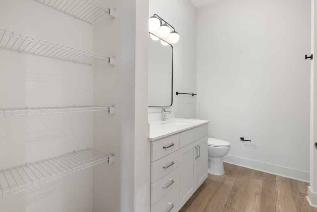 Modern bathroom in a Two Bedroom Apartment with white vanity, mirror, toilet, and open wire shelving on the left; features light wood flooring and neutral walls.