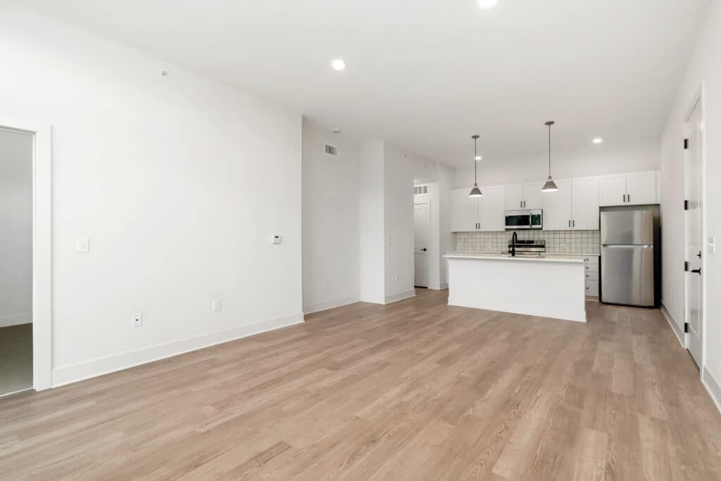 Unfurnished one bedroom apartment with light wood flooring, white walls, an open kitchen featuring white cabinets, stainless steel appliances, and pendant lights. A doorway to a separate room is visible.