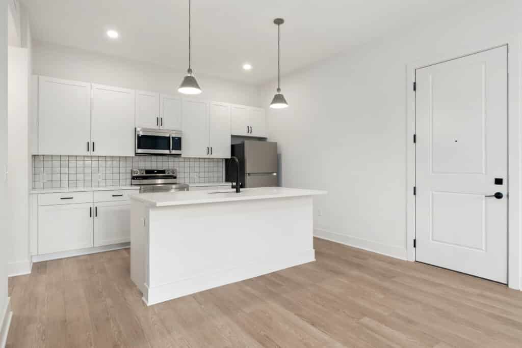 Modern kitchen in a One Bedroom Apartment featuring white cabinets, stainless steel appliances, pendant lights, an island with sink, and light wood flooring.