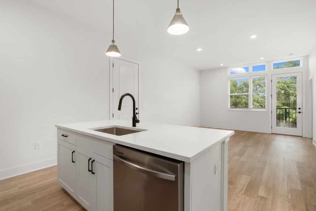 A modern, unfurnished one bedroom apartment features a kitchen and living area with white walls, wood flooring, a central island with a sink, pendant lights, and large windows letting in natural light.