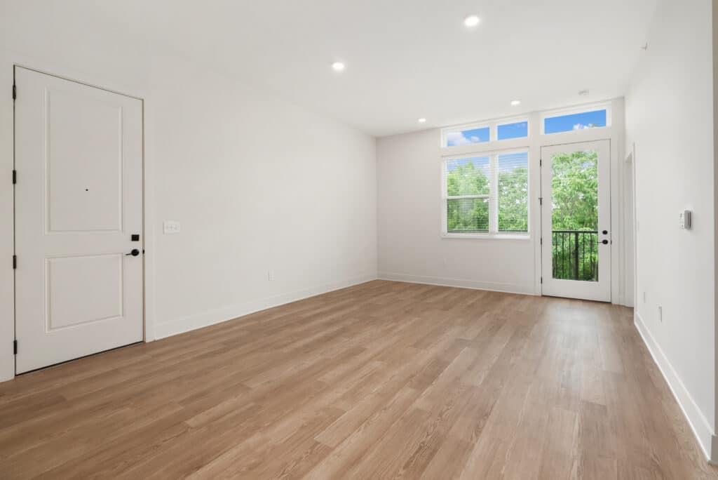 Unfurnished, empty one bedroom apartment with wood flooring, white walls, a closed white door, large window, and glass door leading to a small balcony with outdoor greenery visible.