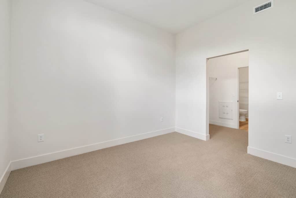 Empty room with beige carpet and white walls in a one bedroom apartment, featuring a small doorway that leads to a washer and dryer area and offers a glimpse of the bathroom.